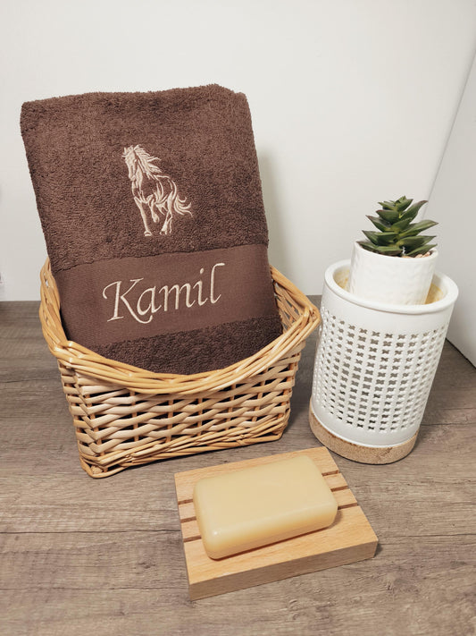 Dunkelbraunes, flauschiges Handtuch in einem Flechtkorb, bestickt mit einem galoppierenden Pferd und dem Namen Kamil in heller Garnfarbe, dekoriert mit Seife und einer kleinen Pflanze.