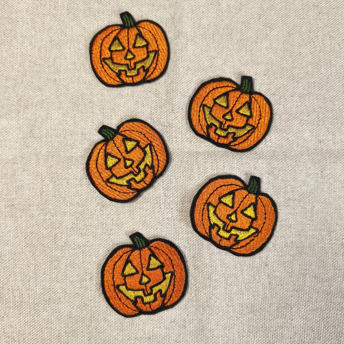 Fünf orangefarbene, bestickte Kürbis-Aufnäher mit gruseligen Jack O'Lantern Gesichtern, flach angeordnet auf einem hellen Leinenstoff.