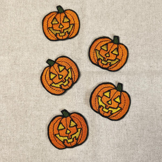 Fünf orangefarbene, bestickte Kürbis-Aufnäher mit gruseligen Jack O'Lantern Gesichtern, flach angeordnet auf einem hellen Leinenstoff.