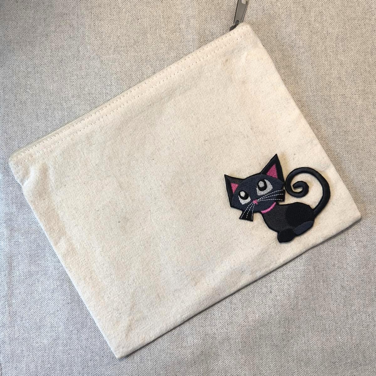 Cremefarbene Canvas-Reißverschlusstasche mit einem schwarzen Katzen-Applikation. Die Katze hat rosa Akzente an Halsband und Ohren sowie einen lockigen Schwanz. Der Reißverschluss ist silberfarben.
