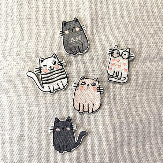 Fünf verschiedene bestickte Katzen-Aufnäher (Patches) zum Aufbügeln, darunter eine Katze mit Brille, eine mit Love-Schriftzug und eine zwinkernde Katze, flach liegend auf Leinenstoff.