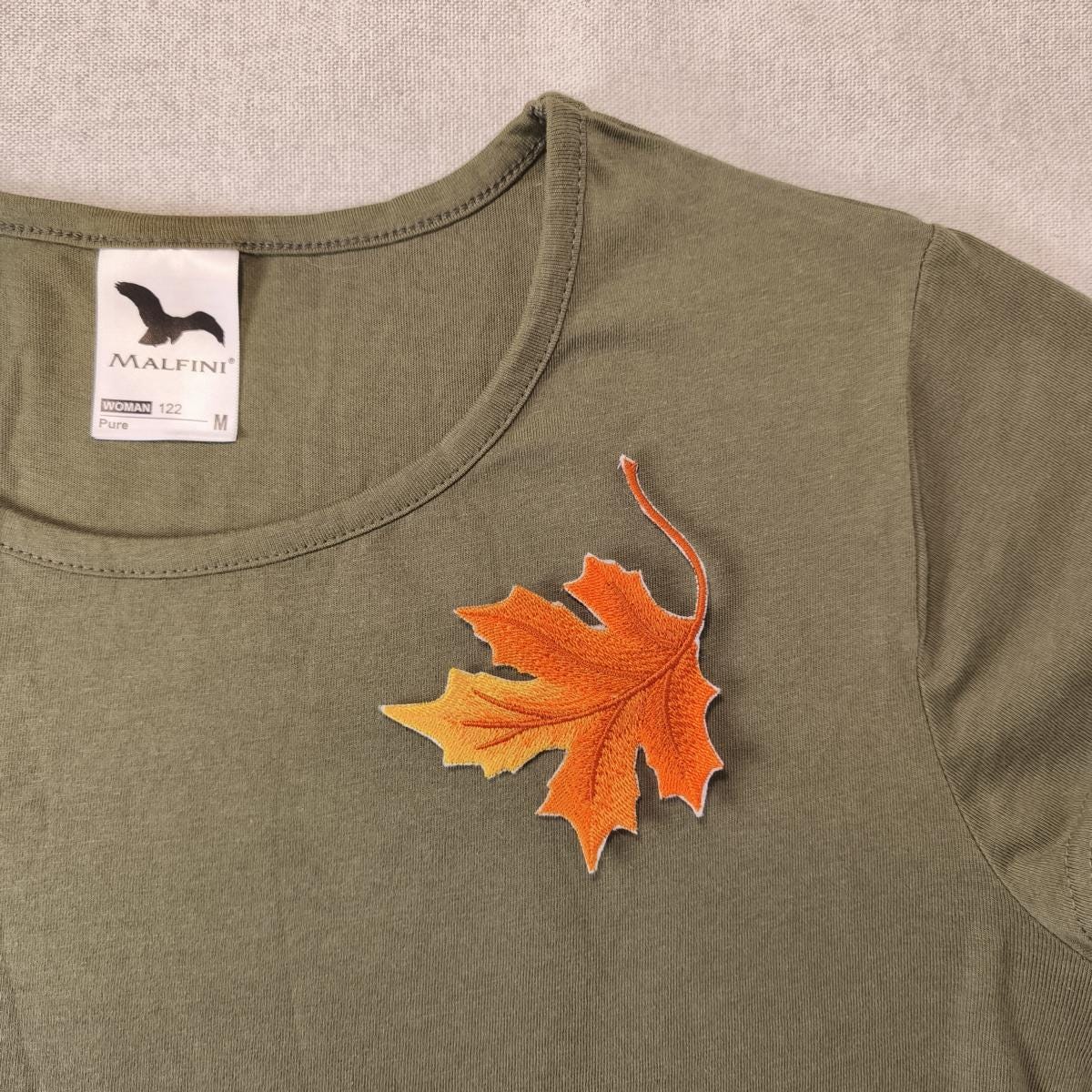 Ein Ahornblatt Aufnäher zum aufbügeln in Herbstfarben auf einem olivgrünen T-Shirt als Beispiel auf einem neutralen Hintergrund
