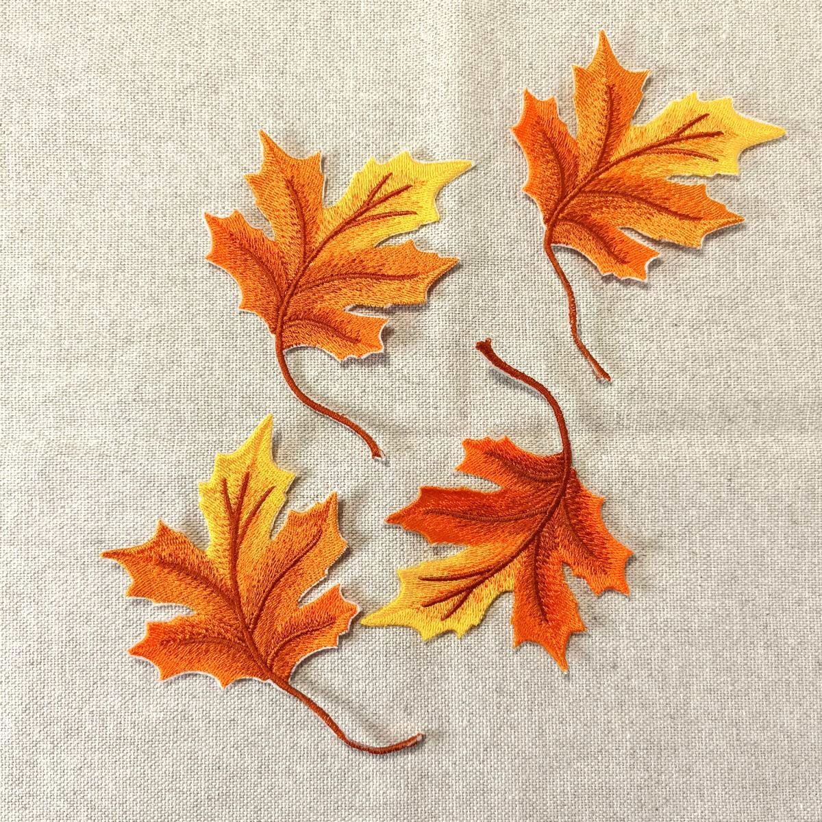 Vier detailreich bestickte Ahornblätter in herbstlichen Orange- und Gelbtönen mit realistischen Blattadern, flach liegend auf einem groben Leinenstoff.