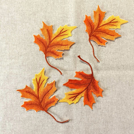 Vier detailreich bestickte Ahornblätter in herbstlichen Orange- und Gelbtönen mit realistischen Blattadern, flach liegend auf einem groben Leinenstoff.