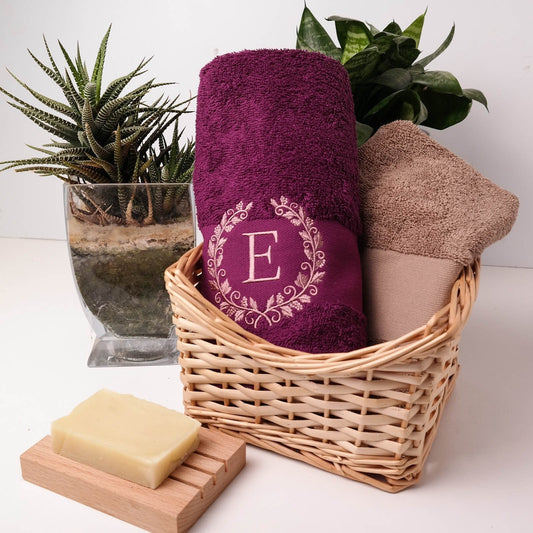 Ein lilafarbenes Badetuch mit einer gestickten Blumenkrone und dem Buchstaben "E" in der Mitte. Das Handtuch liegt in einem Weidenkorb mit einem braunen Handtuch. Ein Stück Seife liegt auf einem Holzseifenhalter.