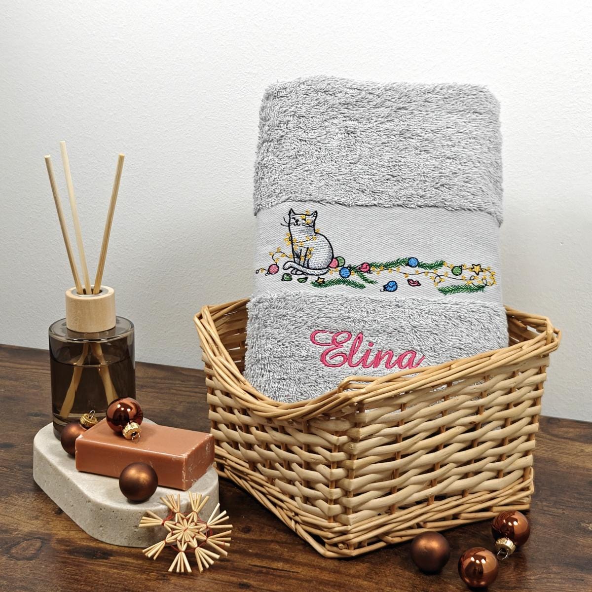 Hellgraues flauschiges Handtuch in einem Weidenkorb, bestickt mit einer sitzenden Katze in einer bunten Lichterkette und dem Namen Elina in pinker Schreibschrift, weihnachtlich dekoriert.
