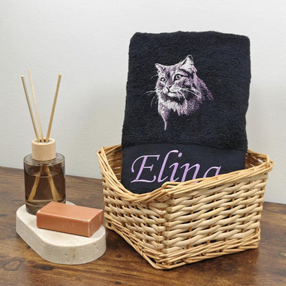 Schwarzes Frotteehandtuch mit einer detaillierten lila-weißen Stickerei eines Langhaarkatzen-Gesichts und dem Namen Elina in violetter Schreibschrift, dekorativ in einem Korb platziert.