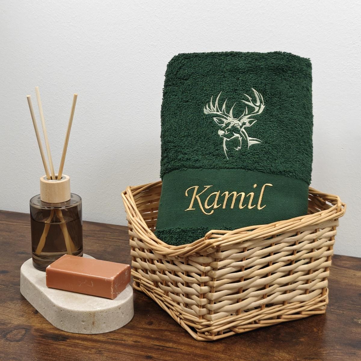 Dunkelgrünes Baumwollhandtuch in einem Weidenkorb, bestickt mit einer weißen Hirschkopf-Silhouette und dem Namen Kamil in goldener Schreibschrift, dekoriert mit Raumduft und Seife.