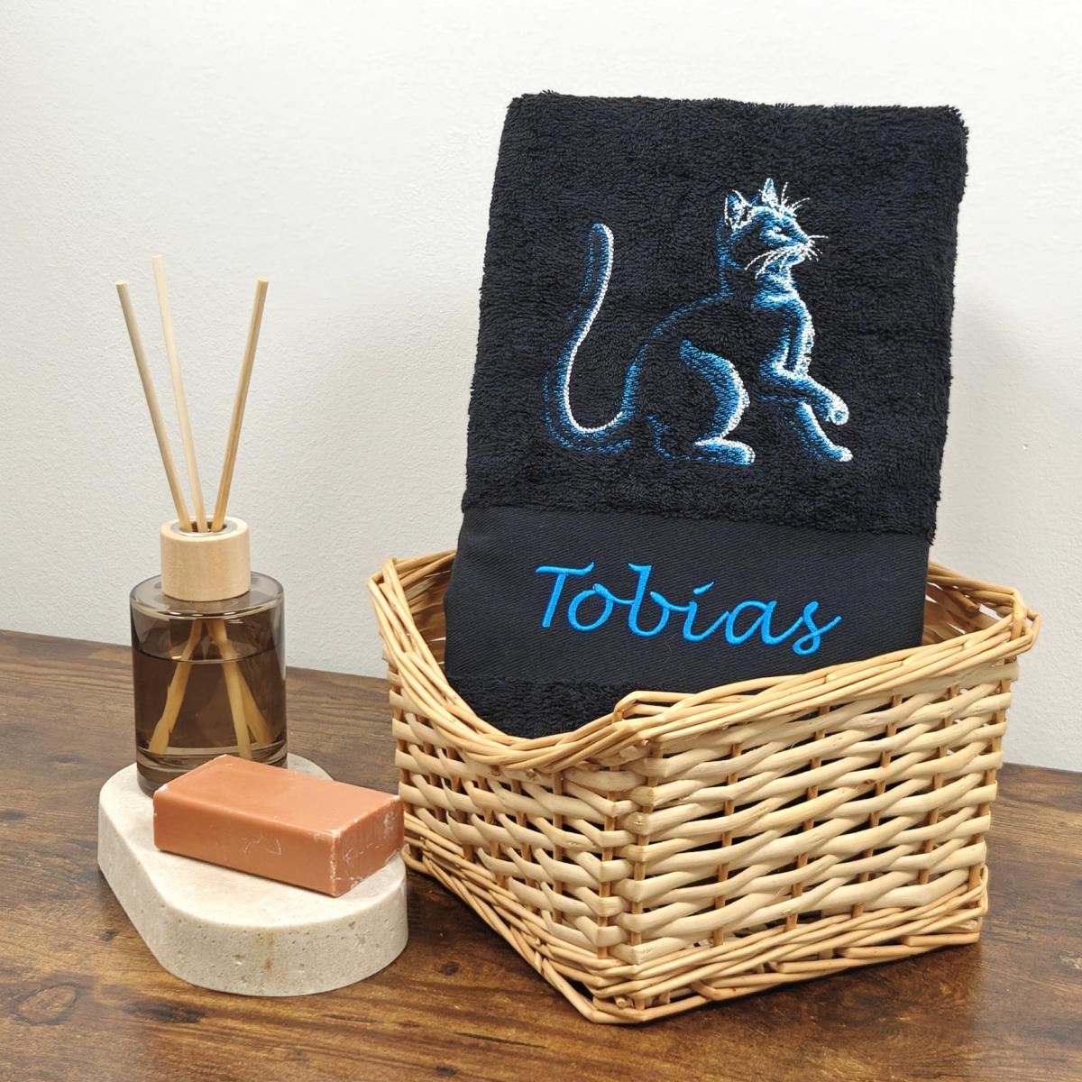 Schwarzes Duschtuch mit einer blau schimmernden Stickerei einer sitzenden Katze und dem Namen Tobias in blauer Schreibschrift, stilvoll arrangiert in einem hellen Flechtkorb.