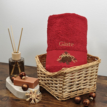 Rotes flauschiges Handtuch in einem Flechtkorb, bestickt mit zwei braunen Tannenzapfen an einer goldenen Schleife und dem Wort Gäste in Gold, dekoriert mit weihnachtlichen Kugeln und Raumduft.