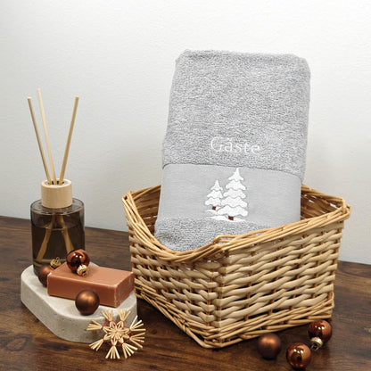 Hellgraues flauschiges Handtuch in einem Weidenkorb, bestickt mit zwei weiß-glitzernden Wintertannen und dem Wort Gäste, weihnachtlich dekoriert mit braunen Christbaumkugeln und Strohsternen.