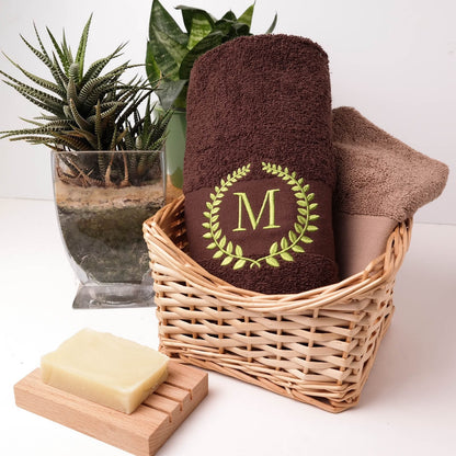 Braunes flauschiges Handtuch mit grüner Bestickung eines Lorbeerkranzes und dem Buchstaben M in einem Weidenkorb neben einer Pflanze und Seife.