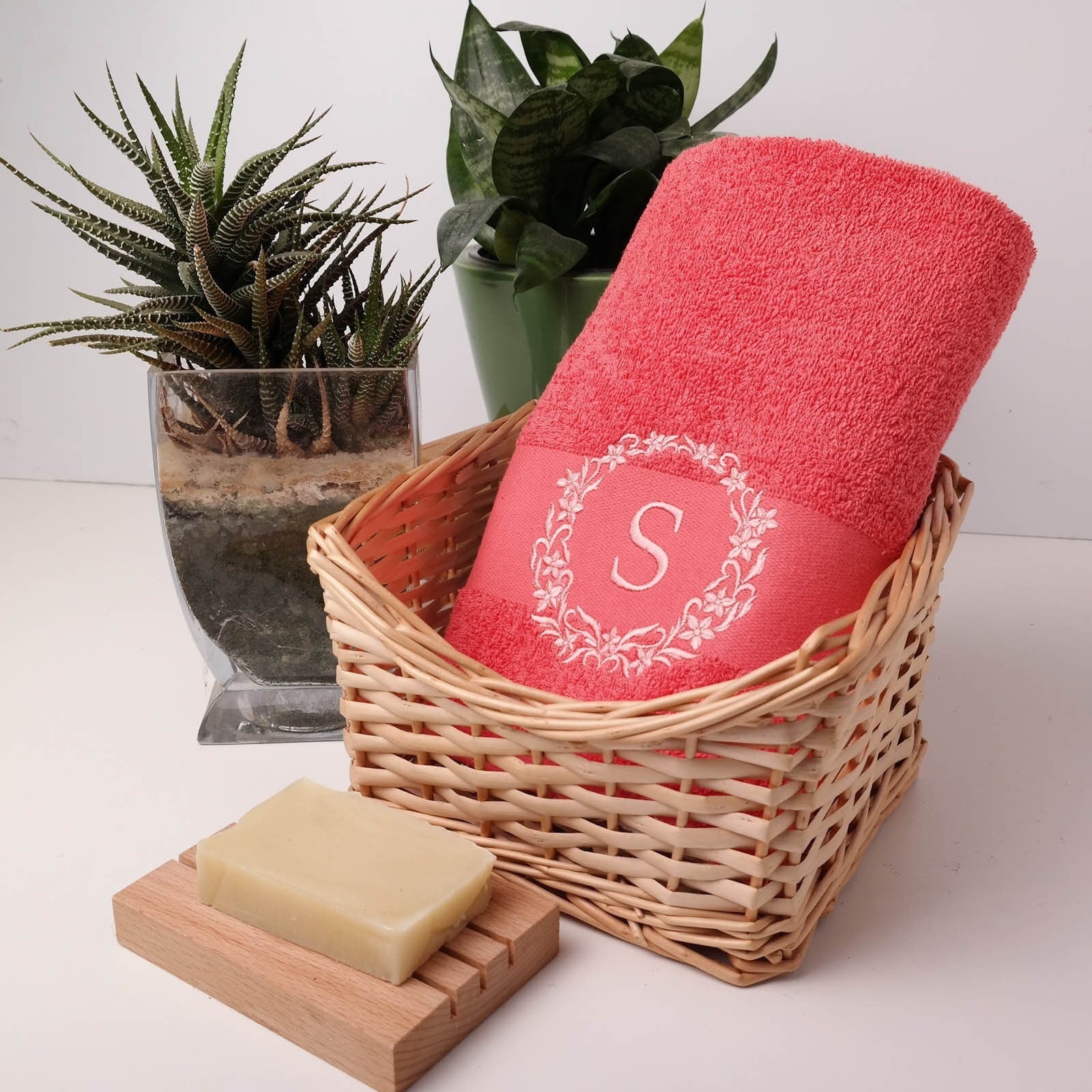 Korallenrotes (Peach) flauschiges Handtuch in einem Weidenkorb, bestickt mit einem weißen Blumenkranz und dem Initial S, dekoriert mit grünen Sukkulenten und Naturseife.